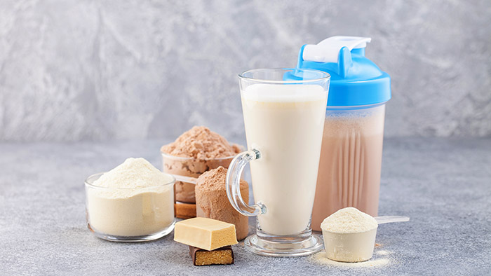 A glass of milk-based protein shake surrounded by scoops and bowls of vanilla and chocolate protein powder, protein bars, and a shaker bottle.